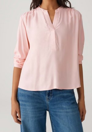 Femme portant un chemisier rose clair à manches longues avec une encolure fendue et un jean bleu, debout devant un fond neutre.