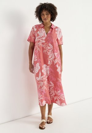 Kobieta z kręconymi włosami w koralowo-różowej, kwiecistej sukience midi z guzikami i białych płaskich sandałach, stojąca przy białej ścianie.