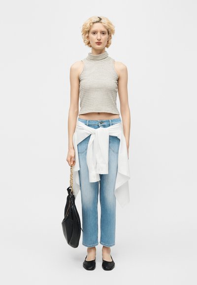 Jeune personne aux cheveux blonds courts et bouclés, portant un crop top rayé sans manches, un jean bleu avec une chemise blanche nouée à la taille, des ballerines noires, et tenant un sac noir.