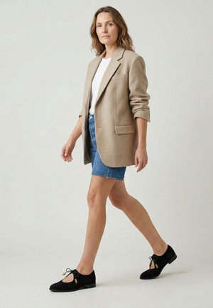 Kvinde går iført beige blazer, hvid top, blå denimshorts og sorte snørestøvler mod en ensartet lys baggrund.
