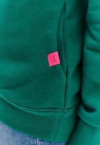 Zelená mikina se žebrovaným lemem a manžetami, která má na postranní kapse růžovou nášivku s grafickým designem. Měkká směs bavlny.