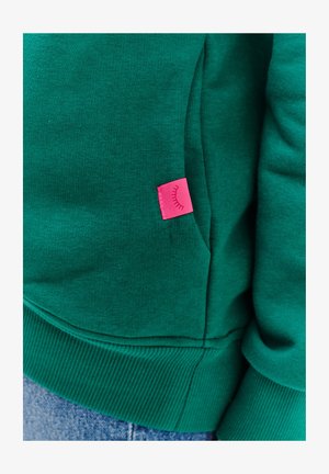 Zielona bluza z ribbowanym hemem i mankietami, z różową metką z graficznym wzorem na bocznej kieszeni. Miękki materiał z mieszanki bawełny.