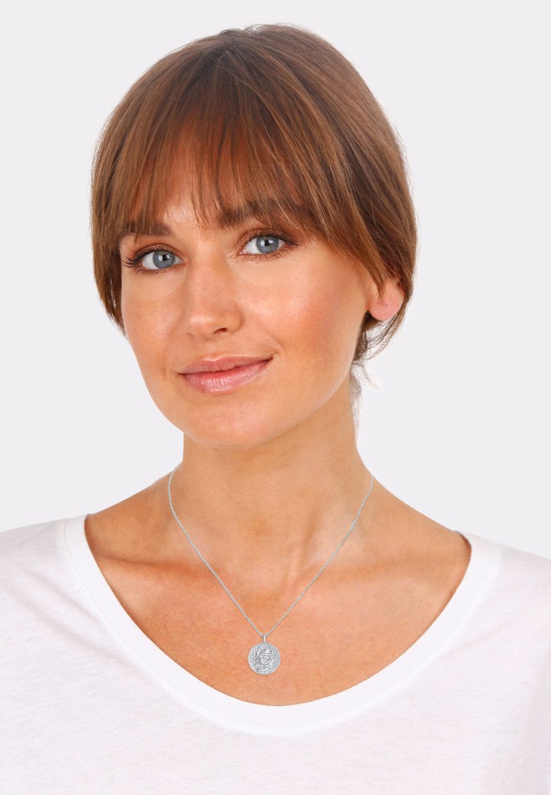 Collana in tono argento con un pendente rotondo e intricato. La persona che la indossa ha una camicia bianca con scollatura a V e capelli castani lunghi fino alle spalle.