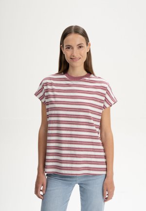 Vrouw met lang bruin haar die lacht, gekleed in een roze-wit gestreept shirt met korte mouwen en lichtblauwe jeans, tegen een effen achtergrond.