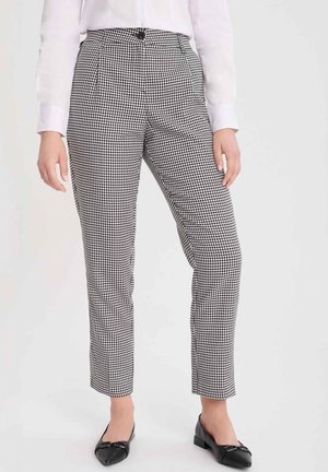 Vrouw draagt zwarte en witte broek met een pied-de-poule-patroon en hoge taille, gecombineerd met een wit overhemd en zwarte platte schoenen.