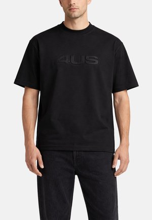Hombre que lleva una camiseta negra lisa de manga corta con el logo en relieve "4US" en el pecho y vaqueros negros, de pie frente a un fondo blanco.