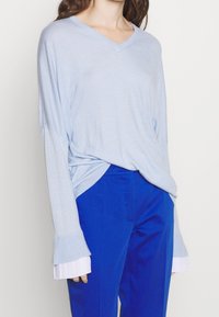 Ljusblå, långärmad V-ringad tröja med ribbade muddar, lagd över vit plisserad väv, i kombination med klara blå byxor.