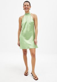 Satin miniklänning i ljusgrön med halterneck-design, slät textur, med en avslappnad passform och utsvängd nederkant. Bärs med bruna flip-flop-sandaler.