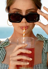 Zwarte oversized zonnebril, gestreepte rietje in een glas met een rode drank, een geribde glazen vasthoudend, groen-wit geblokte jurk met gestrikte bandjes.