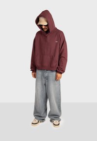 Felpa cappuccio color marrone bordeaux con tasca frontale e piccolo logo. Indossata con jeans blu larghi e sneakers crema con dettagli neri.