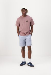 Helles rosa Baumwoll-T-Shirt mit einem kleinen Bergmotiv, kombiniert mit blau gestreiften Shorts. Ergänzungen: weißer Bucket Hat, marineblaue Sneaker.