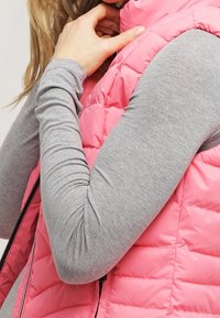 Rosa puffig väst med diagonalt quiltat mönster, bärs över en långärmad grå topp. Har en hög krage och framzipper.