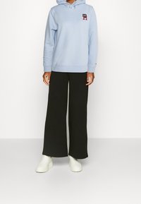 Jasnoniebieska bluza z kapturem z przednią kieszenią i ściągaczem, w połączeniu z luźnymi czarnymi spodniami i białymi tenisówkami. Bluza posiada logo na piersi.