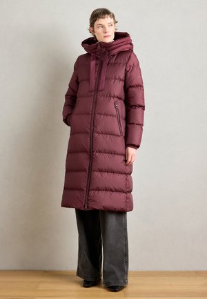 Vrouw in een lange, gewatteerde wijnrode winterjas met capuchon, gekleed in een brede, donkere grijze broek, staande tegen een effen lichte muur.
