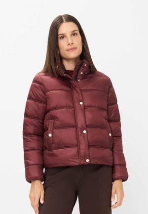 Frau mit glattem braunem Haar, die eine burgunderrote Steppjacke mit Druckknöpfen und dunkelbraune Hose vor einem schlichten weißen Hintergrund trägt.