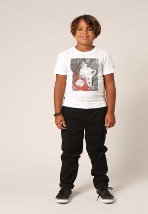 Witte T-shirt met een afbeelding van een rode elektrische gitaar, gecombineerd met zwarte cargobroeken en zwarte sneakers. Bruine armband om de pols.