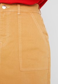 Pantalons en denim beige avec une texture lisse, dotés de coutures blanches, d'une poche avant et d'une fermeture à bouton en métal au niveau de la taille.