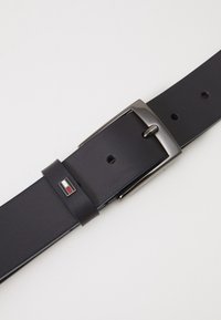 Ceinture en cuir noir avec une texture lisse, boucle rectangulaire en argent, et un accent de logo en rouge, blanc et bleu sur une boucle près de la boucle.