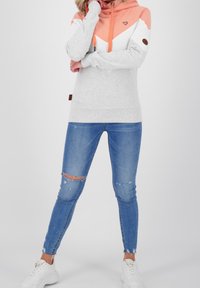Felpa grigia con design a chevron arancione e bianco, dotata di tasca a marsupio e lacci coordinati. Indossata con jeans blu strappati e sneakers bianche.