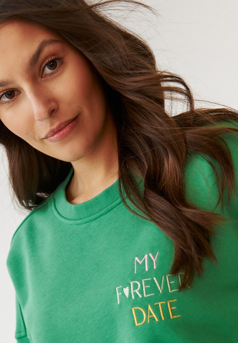 Zielona bluza z okrągłym dekoltem, na której znajduje się wielokolorowy haftowany napis: "MOJA WIECZNA RANDKA". Materiał wydaje się miękki i teksturowany.