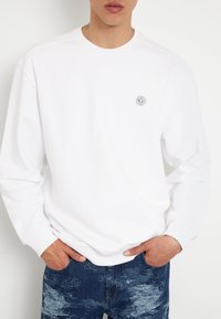 Valge pusakun, millel on ümar kaelus ja soonikkoes varrukad. Esiküljel on väike ümmargune logo. Kombineeritud siniste kirjustruktuursete teksadega.