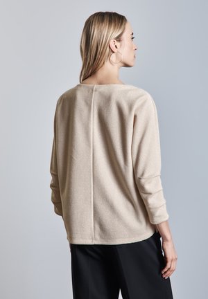 Pull beige texturé avec une coupe ample, des manches trois-quarts et une couture arrière droite, associé à un pantalon noir.