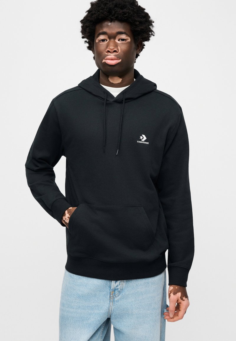 Sweat à capuche en polaire noire avec poche kangourou et cordon de serrage, arborant un petit logo Converse blanc sur la poitrine. Porté avec un jean bleu clair.
