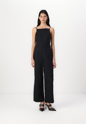 Black sleeveless jumpsuit with a square neckline, tailored fit, wide legs, and structured waist. Accompanied by black high-heeled shoes with buckles.