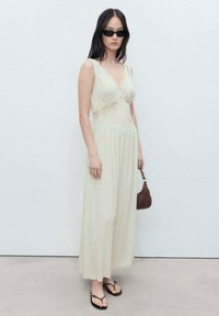 Lightly patterned, pale green maxi dress with a V-neck and lace accents, paired with black sunglasses and a brown handbag. Flip-flops complete the look.