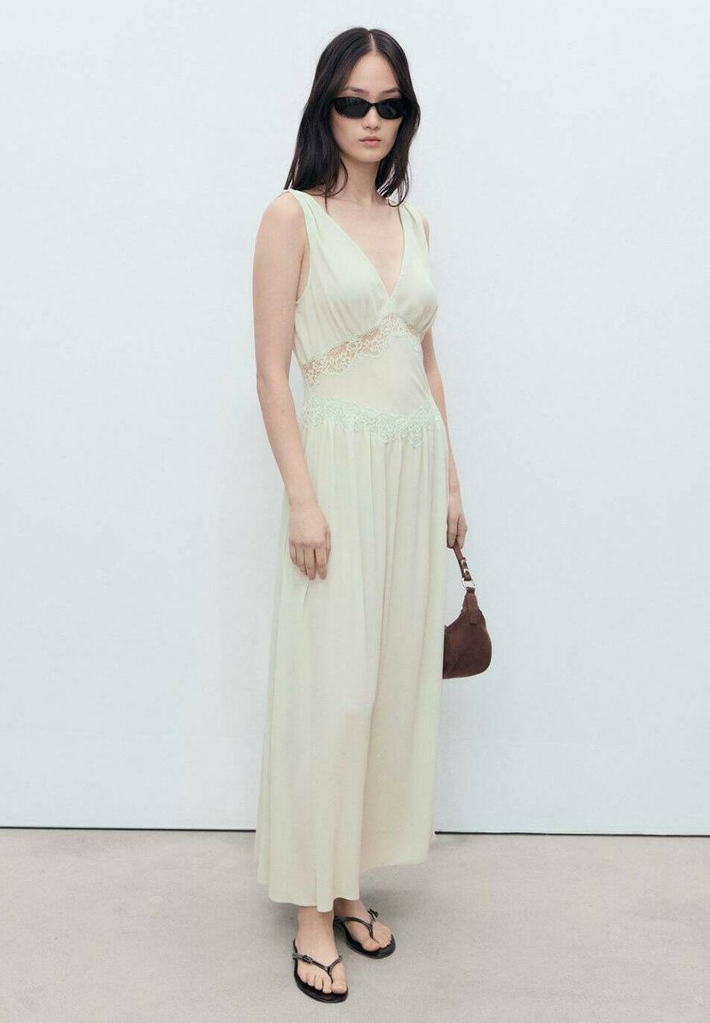 Lightly patterned, pale green maxi dress with a V-neck and lace accents, paired with black sunglasses and a brown handbag. Flip-flops complete the look.