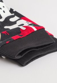 Sweaterærme af mønstret stof med sort bund, røde accenter og hvide ko-motiver; har en ribbet sort manchet for tekstur.