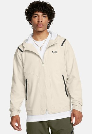 Giacca zip-up color beige chiaro con cappuccio, dettagli neri, due tasche laterali e una texture liscia. Logo visibile sulla parte sinistra del petto.