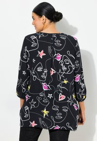 Schwarze Bluse mit Umrissen von Gesichtern in Weiß und Akzenten von pinken, gelben und roten Sternen. Verfügt über Dreiviertelärmel und einen Rückenfalte.