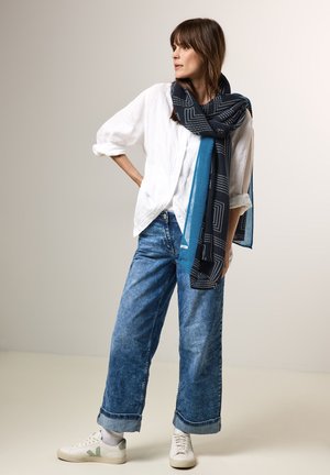 Denim breddebenede jeans, hvid linned skjorte og et mønstret marineblåt tørklæde med geometriske designs. Hvide sneakers med grønne detaljer.