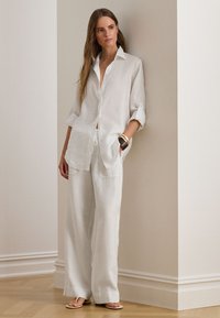 Fehér lenvászon ing felhajtott ujjakkal, laza fehér lenvászon nadrággal párosítva. Jellegzetes részletek a elülső zsebek és a kényelmes, bő szabás.