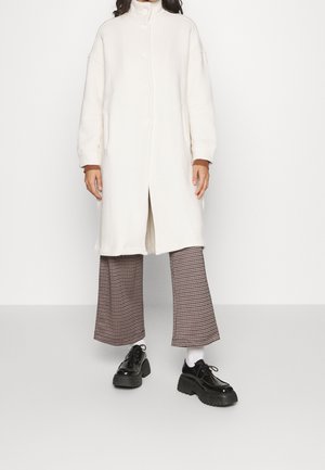 Personne portant un long manteau blanc boutonné, un pantalon large à carreaux marron, des chaussettes blanches et des chaussures compensées noires sur fond blanc.