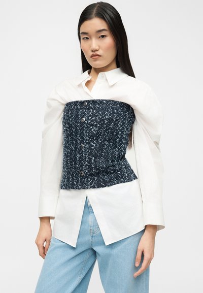 Jeune femme portant une chemise blanche boutonnée sous un corset texturé bleu marine et un jean bleu clair, debout devant un fond uni.