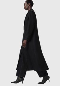 Abrigo largo negro con un diseño estructurado, aberturas laterales y un ajuste entallado. Llevado sobre pantalones oscuros y botas de tacón negro puntiagudas.