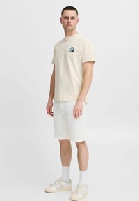 Beige cotton t-shirt with small wave graphic on left chest, paired with white linen shorts and beige sneakers, against a plain background.