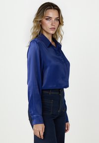 Szatynowa niebieska bluzka z kołnierzykiem na guziki, długimi rękawami i gładką teksturą, w zestawieniu z ciemnymi dżinsami z wysokim stanem.