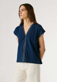 Kobieta o długich brązowych włosach, ubrana w granatową bluzkę z krótkim rękawem z ozdobnym wykończeniem, w połączeniu z kremowymi spodniami, patrząca w lewo.