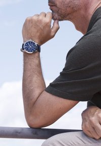 Okrągły niebieski zegarek chronograf z srebrnymi akcentami i granatowym skórzanym paskiem, umieszczony na nadgarstku opartym o metalową balustradę.