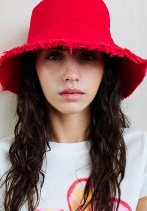 Femme aux longs cheveux bouclés foncés portant un chapeau bob rouge vif et une chemise blanche avec un motif circulaire coloré.