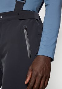 Schwarze wasserdichte Hosen mit einer Reißverschlusstasche und verstellbaren Trägern, kombiniert mit einem hellblauen Langarmshirt, das eine detaillierte Textur zeigt.