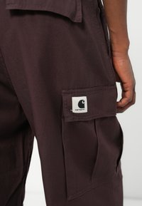 Pantaloni cargo viola scuro con molteplici tasche, una consistenza morbida e una patch con il marchio ben visibile sul lato.
