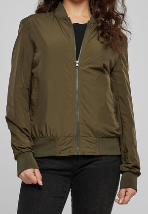 Mujer con chaqueta bomber verde oliva con puños y cinturilla acanalados, camiseta negra debajo y pantalones negros.