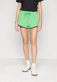 Gröna bomullsshorts med dragsko i midjan och marinblå kant, med en logotyp på vänster sida. Matcher med vita sneakers och strumpor.