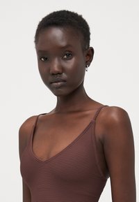 Débardeur à rayures marron avec des fines bretelles et un décolleté en V plongeant, en tissu lisse, de design ajusté. Boucle d'oreille en argent visible.