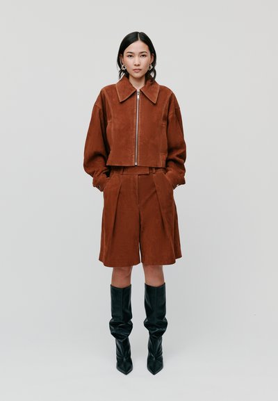 Braune Wildleder-Top-Jacke mit breitem Kragen, kombiniert mit passenden, gefältelten Shorts. Das Model trägt kniehohe, lässige, schwarze Stiefeletten mit spitzer Schuhspitze.
