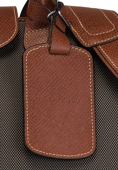 Braunes Lederetikett mit strukturierten Oberflächen, mit eingeprägtem "LONGCHAMP"-Logo, befestigt an einem dunkel gemusterten Stoff. Doppelte Lederklappendetails.
